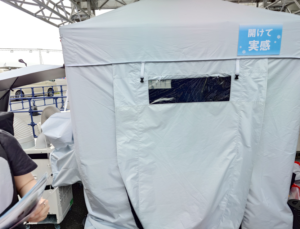 猛暑対策展で展示された山善株式会社の「冷却シェルター」。白いテント型の外観で、「開けて実感」と書かれた青いポップが貼られ、冷却用ダクトなどの設備が接続されている