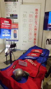 猛暑対策展に出展された「熱中症救護袋」のブース。「熱中症になった時に使う商品です。『熱中症救護袋』」と書かれた立て看板と、赤と青を基調とした救護用の搬送・冷却バッグの製品実物が展示されている。