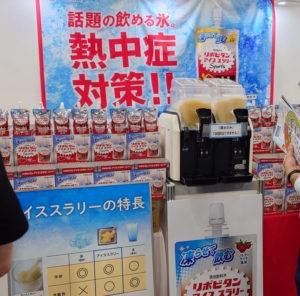Promotional booth featuring a large blue banner with red Japanese text about heatstroke prevention, two yellow slush machines, and rows of drink cartons displayed on the counter behind them.