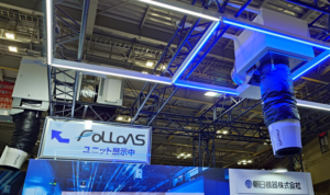 猛暑対策展で出展された朝日機器株式会社の「FOLLOAS(フォロアス)」ユニット展示。「FOLLOAS ユニット展示中」の看板とともに、天井のトラスフレームに取り付けられた青いLED照明と大型のスポット空調・送風ダクト設備が写されている。