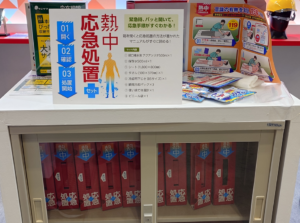 猛暑対策展で展示された「熱中症応急処置セット」。緊急時の手順が書かれた案内板と同梱アイテム(アクアソリタ、冷却パック、タオル等)が置かれ、下の棚には専用の赤い収納ボックスが並んでいる。
