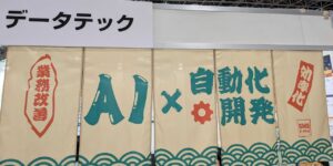At a trade show booth, beige fabric banners display teal text reading 'AI × 自動化開発' with red vertical stamps saying '業務改善' and '効率化', and a teal wave pattern along the bottom.